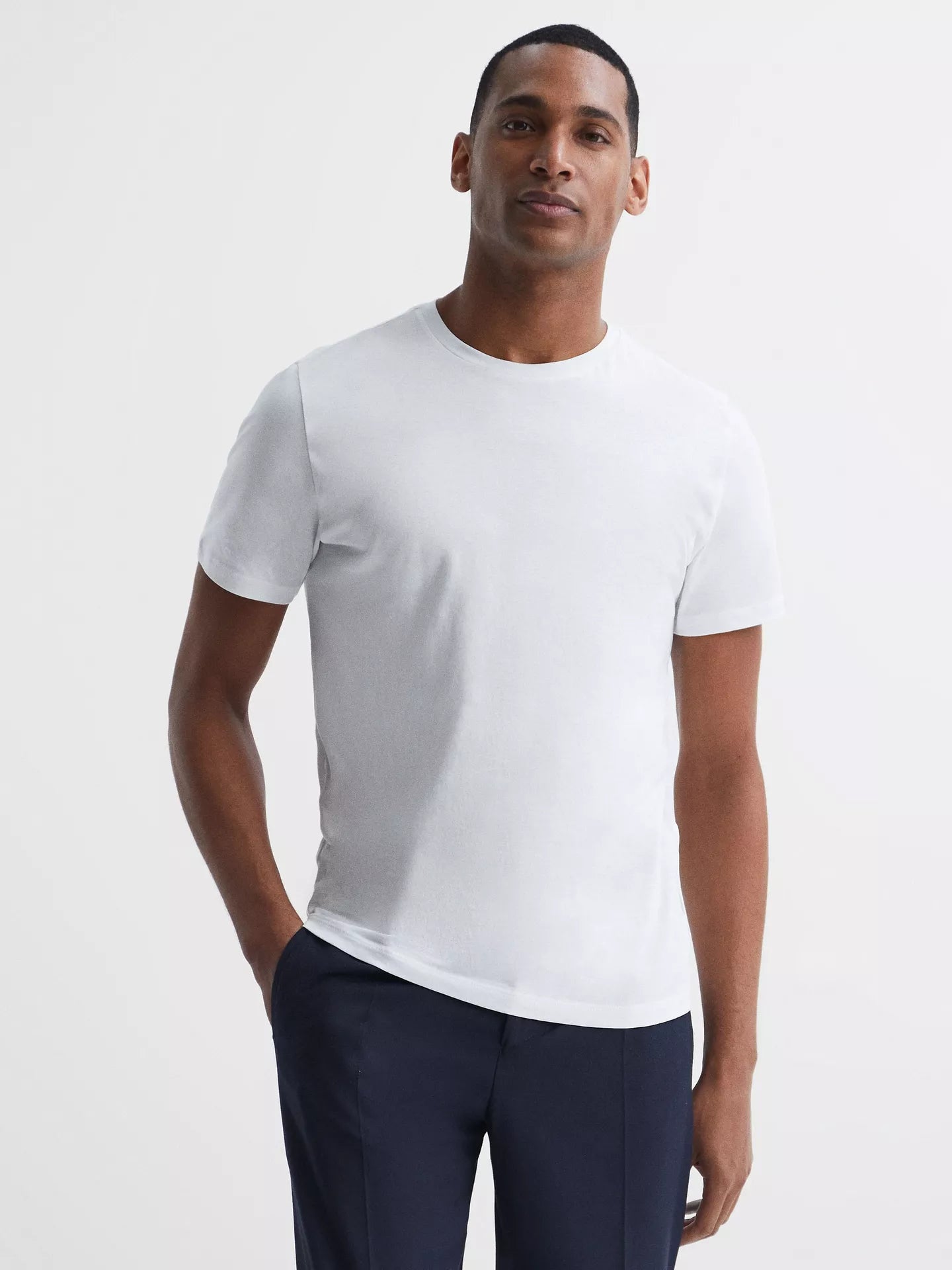 A man wearing a plain white short-sleeve crewneck t-shirt and navy blue trousers, standing against a light gray studio background.