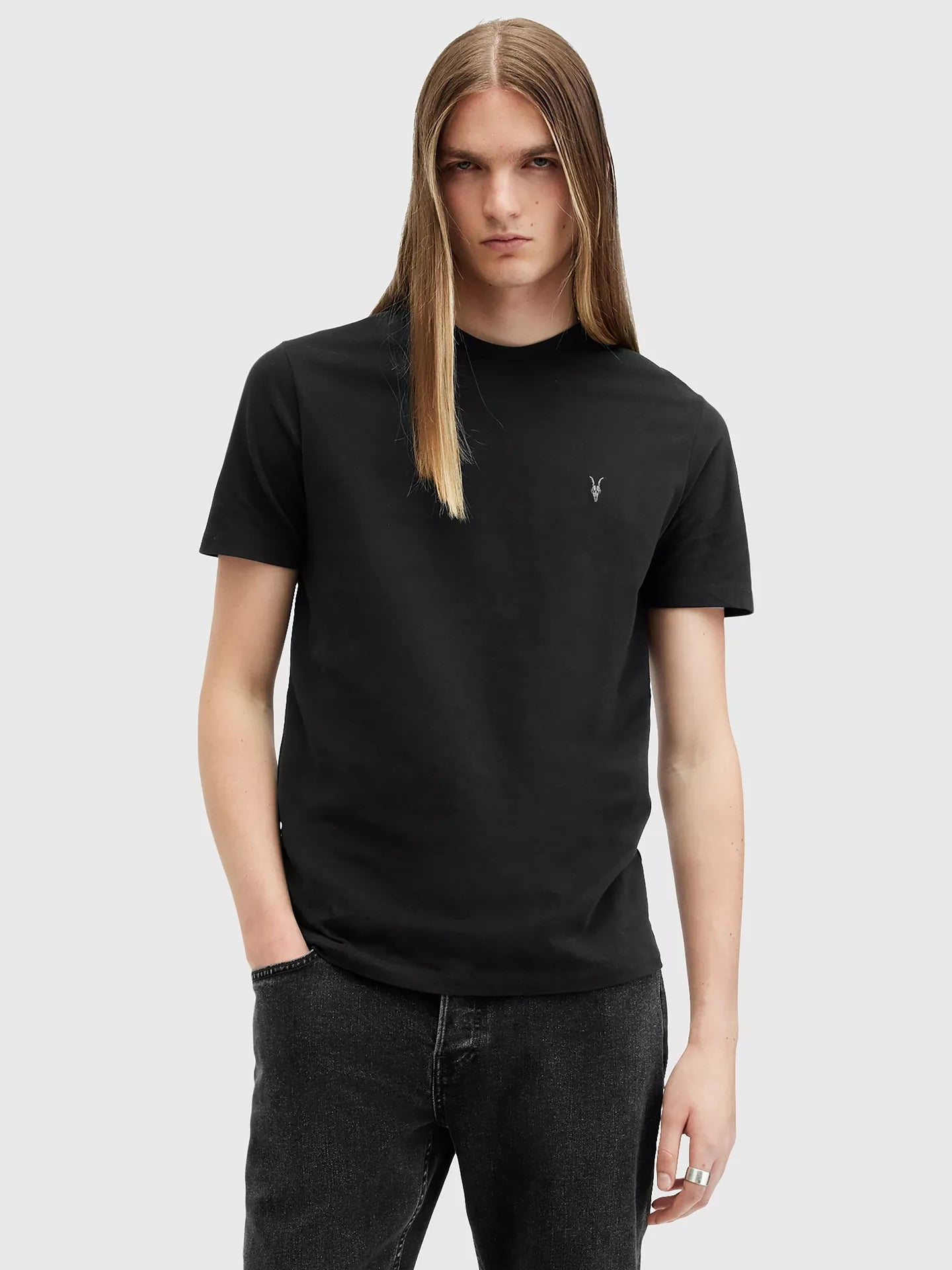 A male model with long blonde hair wearing a black short-sleeve crewneck t-shirt with a small embroidered logo on the left chest, paired with dark gray jeans, against a light gray background.