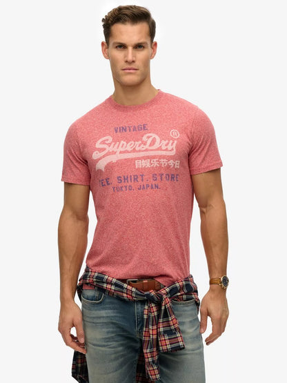 A man wearing a heathered pink Superdry graphic t-shirt with a red and black plaid shirt tied around his waist, paired with blue jeans, against a light gray background.