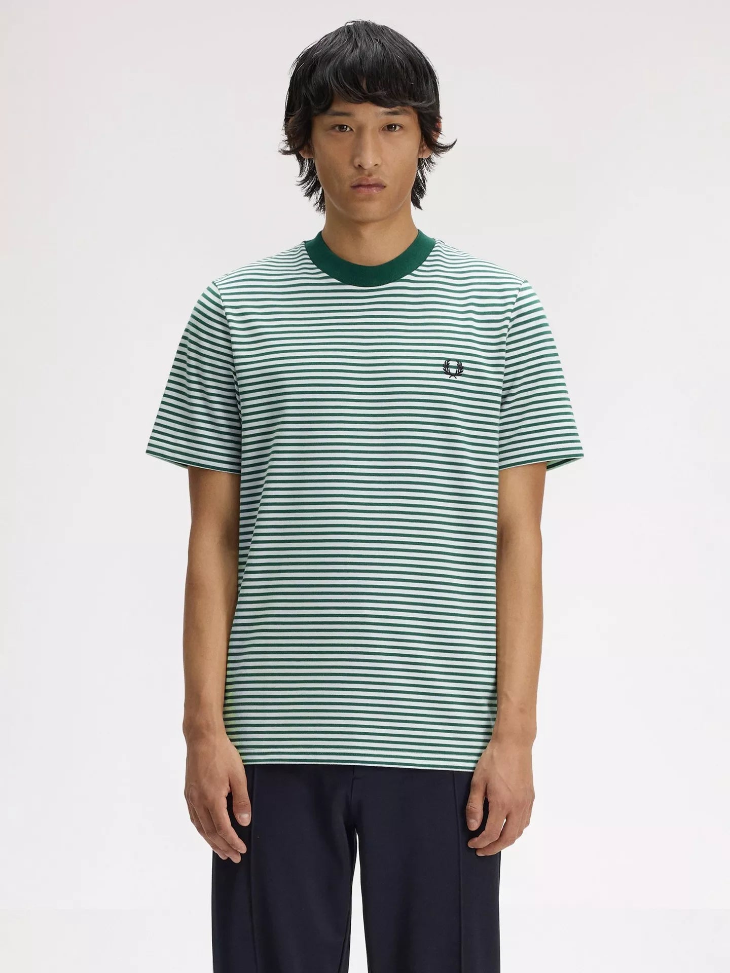 A model wearing a green and white horizontally striped short-sleeve t-shirt with a dark green crew neck and a small embroidered logo on the chest, paired with dark navy pants, against a plain light gray background.