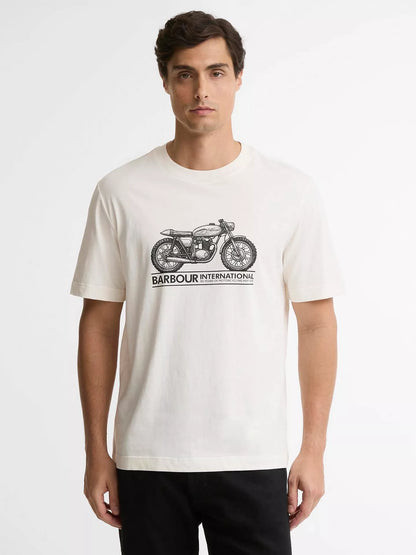 A man wearing a cream-colored short-sleeve t-shirt with a black graphic of a vintage motorcycle and the text 'BARBOUR INTERNATIONAL' printed on the chest, paired with black pants, against a plain light gray background.