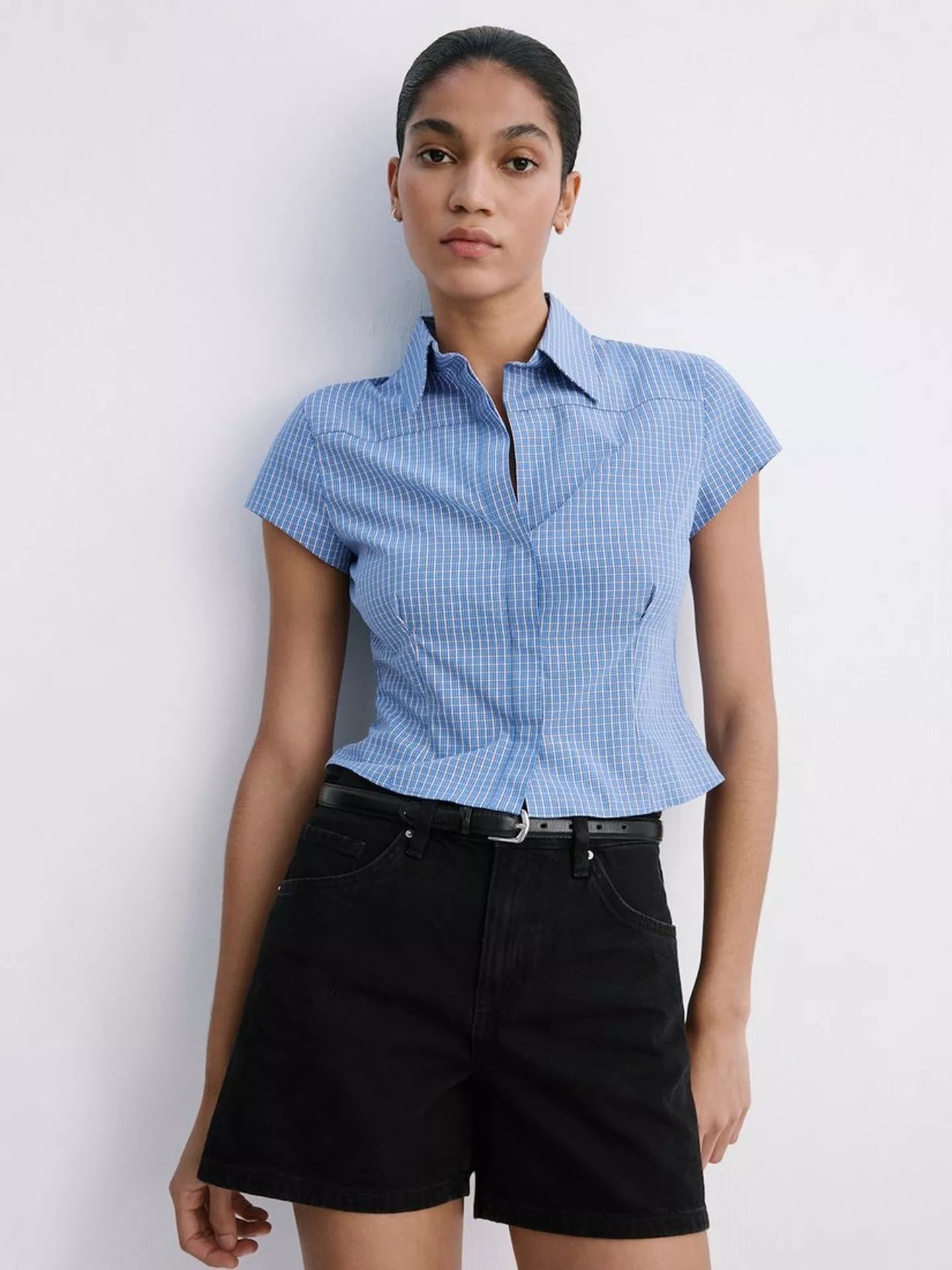 A woman modeling a light blue and white gingham short-sleeve button-up shirt paired with black shorts, standing against a plain white wall.