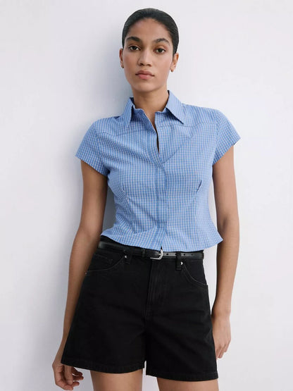 A woman modeling a light blue and white gingham short-sleeve button-up shirt paired with black shorts, standing against a plain white wall.