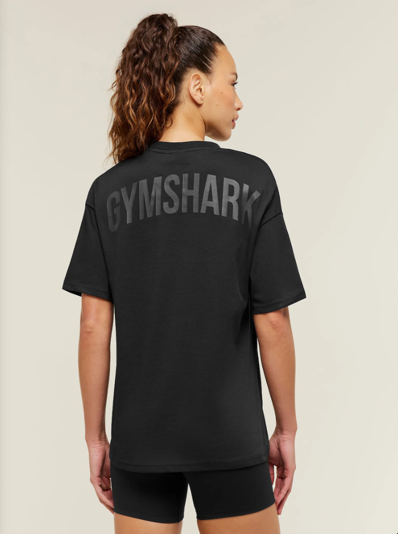 A woman shown from the back wearing a black short-sleeve t-shirt with 'GYMSHARK' printed in large gray letters across the upper back, paired with black shorts against a light beige studio background.
