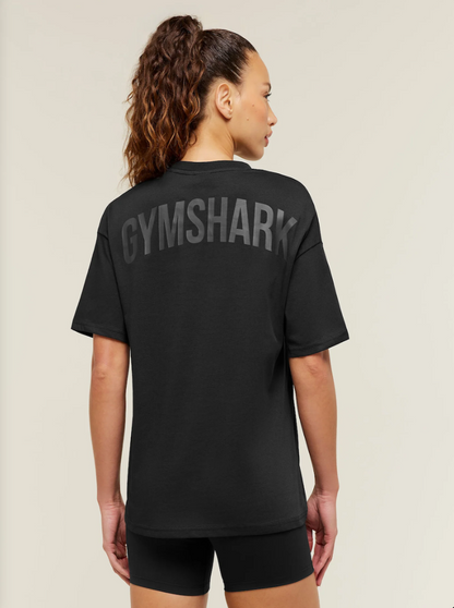 A woman shown from the back wearing a black short-sleeve t-shirt with 'GYMSHARK' printed in large gray letters across the upper back, paired with black shorts against a light beige studio background.