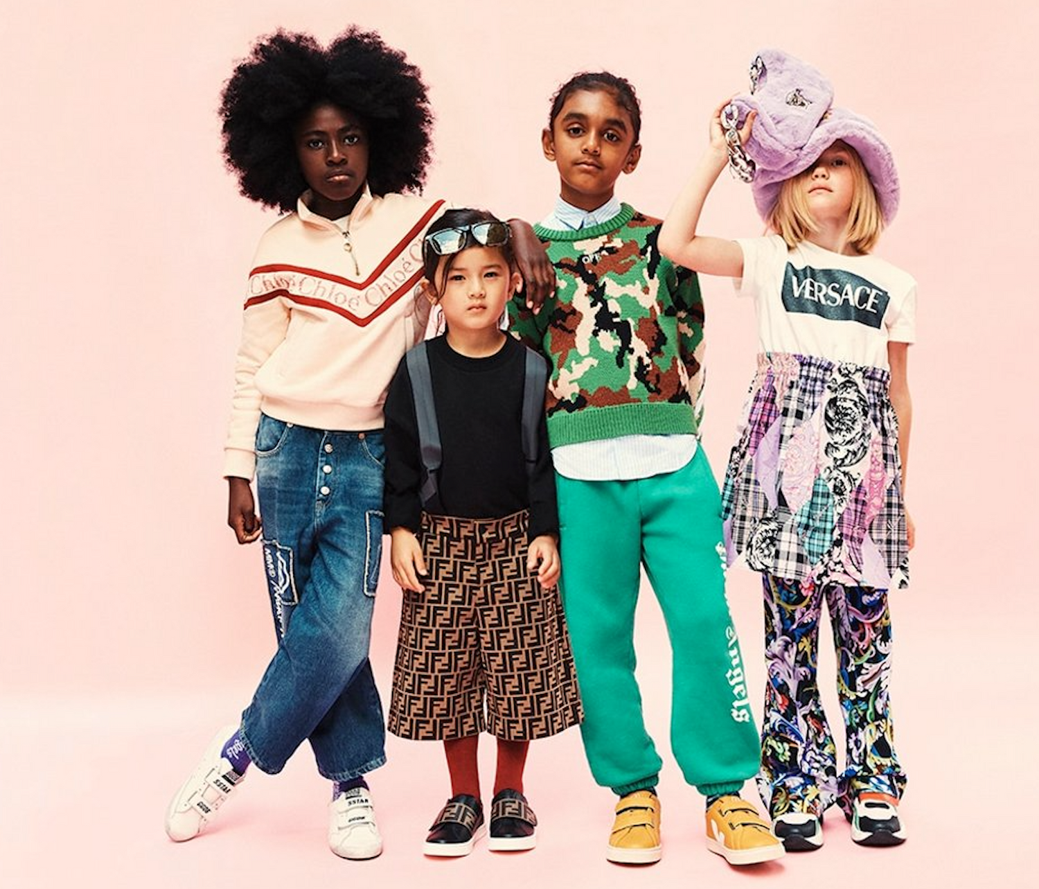 Four children modeling Versace clothing against a solid light pink studio background. The outfits include a cream zip-up hoodie with red stripes, blue jeans, a green camouflage sweater, bright green sweatpants, a white graphic t-shirt, and a purple wide-brim hat.
