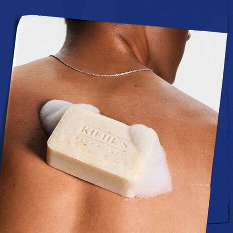 A close-up of a person's bare upper back with a bar of Kiehl's Since 1966 body soap resting on the skin, surrounded by white lather foam, set against a dark blue graphic background.